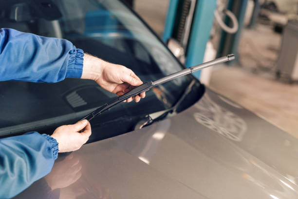 A person wearing a blue jacket holds a detached windshield wiper in their hand over the hood of a car.
